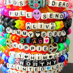 May include: A stack of colorful beaded bracelets with various phrases and charms. The bracelets feature white letter beads spelling out words like "ZEDS DEAD," "FULL SEND," and "WORTHY." Charms include a skull, alien, and peace sign.