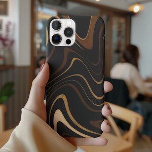 Puede incluir: Funda de teléfono negra con un diseño ondulado en tonos marrón y dorado. La funda tiene un acabado brillante y está diseñada para un teléfono inteligente. La funda del teléfono está en la mano de una persona.