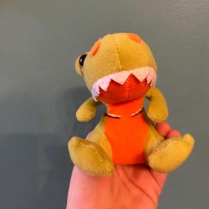 May include: A small, plush dinosaur toy in shades of yellow, orange, and white. The dinosaur has a yellow body, orange belly, and white teeth. It has orange spots on its head and black eyes. The toy is soft and cuddly.
