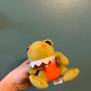 May include: A plush toy frog in shades of yellow and orange. The frog has large black eyes, orange accents, and a white jagged mouth. It is wearing an orange outfit. The toy is soft and cuddly.