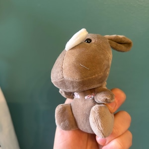 May include: A brown plush toy shaped like a deer with a white patch on its head. The toy has small, dark eyes and a stitched mouth. The deer is holding its arms up and has small, rounded ears. The toy is made of soft, plush material.