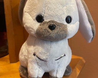 May include: A plush bunny toy in soft white and gray. The bunny has large black eyes, a gray nose and ears, and embroidered details. The toy is sitting on a wooden surface. A cute and cuddly stuffed animal.