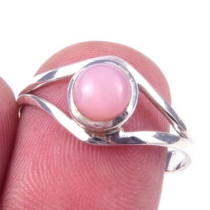 May include: A silver ring with a round, light pink gemstone. The ring's band splits into two curving lines that frame the gemstone. The gemstone is set in a circular bezel.