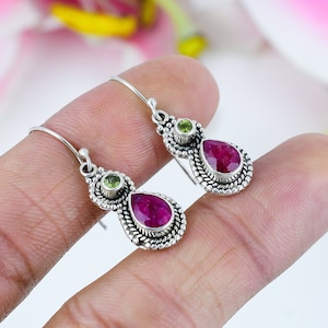 May include: A pair of silver earrings featuring a teardrop-shaped, deep red gemstone, accented with a smaller, round, light green stone. The earrings have a detailed, textured border and a simple hook closure.