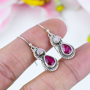 May include: Silver earrings with a teardrop-shaped, faceted, red gemstone and a small, round, pink gemstone. The earrings have a decorative, beaded border and a simple hook closure. The earrings are displayed against a soft, blurred background.