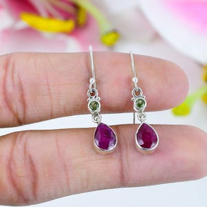 May include: A pair of silver drop earrings featuring a teardrop-shaped, deep red gemstone and a small, round, green gemstone. The earrings are held up by a silver hook.
