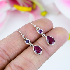 Può includere: Un paio di orecchini pendenti in argento con una piccola pietra di ametista rotonda viola incastonata sopra una gemma di rubino rosa scuro a forma di pera. Gli orecchini sono sospesi a semplici ganci in argento con finitura lucida.