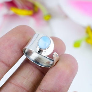 Könnte beinhalten: Ein breiter, polierter Silberring mit einem einzelnen, glatten, ovalen hellblauen Stein in der Mitte. Die Ringschiene ist hinten offen, was eine Anpassung ermöglicht, und hat eine reflektierende, metallische Oberfläche.