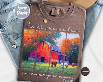 Könnte beinhalten: Espressofarbenes T-Shirt mit einer farbenfrohen Scheunen-Szene und dem Text "I will praise him in every season" und "Psalm 146:2". Das Shirt wird mit einer blauen Jeans und einer goldenen Halskette präsentiert.