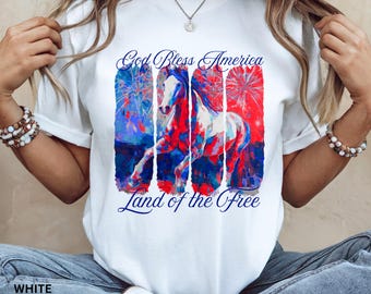 May include: White t-shirt featuring a patriotic design with a horse and fireworks in red, white, and blue. The text "God Bless America" and "Land of the Free" are printed above and below the graphic. The shirt is a Bella Canvas 3001.