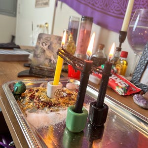 Può includere: Un vassoio d'argento con candele accese in portacandele in ceramica, circondato da fiori secchi, sale e cristalli. Una candela gialla, una candela nera e una candela rossa sono accese. È visibile una carta dei tarocchi con le parole "Acque sacre".