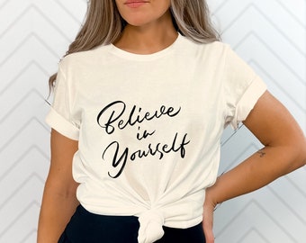 May include: A cream-coloured short-sleeve t-shirt featuring the black cursive text "Believe in Yourself" printed on the front. The shirt has a crew neckline and is tied in a knot at the bottom hem, showcasing a casual and simple design.