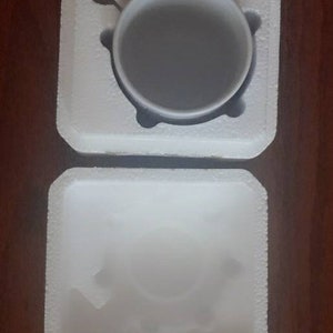 May include: A white ceramic mug with a handle, sitting in a white styrofoam packaging.