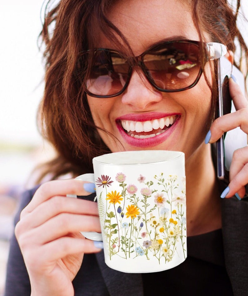 May include: White ceramic mug with a floral design featuring pressed flowers in shades of yellow, pink, blue, and purple. The flowers are arranged vertically on the mug.