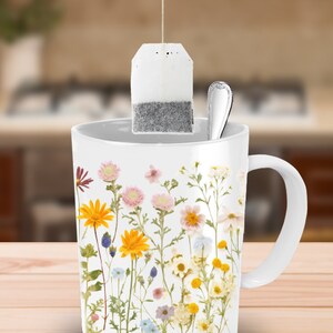 May include: White ceramic mug with a floral design featuring pressed flowers in shades of yellow, pink, white, and purple. A tea bag hangs from the rim of the mug.