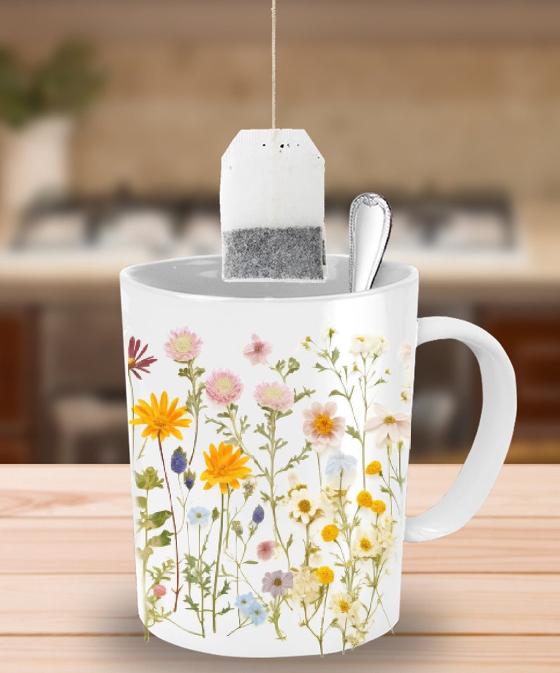 May include: White ceramic mug with a floral design featuring pressed flowers in shades of yellow, pink, white, and purple. A tea bag hangs from the rim of the mug.