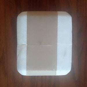 May include: A white foam box with a brown paper tape covering the center. The box is square and has rounded corners.