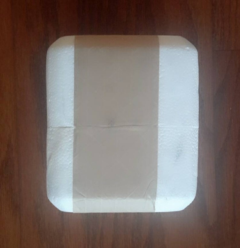 May include: A white foam box with a brown paper tape covering the center. The box is square and has rounded corners.