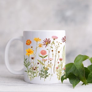 May include: White ceramic mug with a floral design featuring pressed flowers in shades of pink, yellow, and orange. The flowers are arranged in a vertical line on the mug.