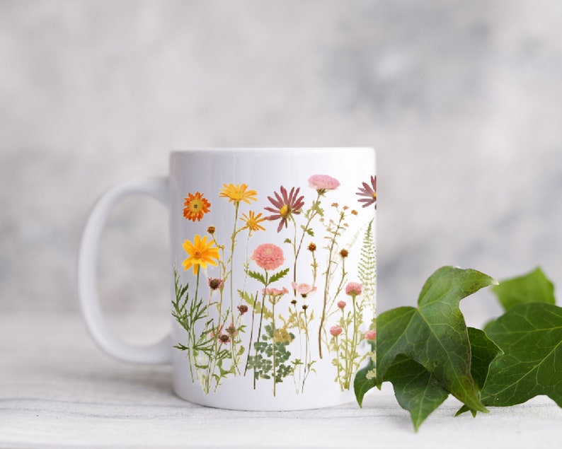 May include: White ceramic mug with a floral design featuring pressed flowers in shades of pink, yellow, and orange. The flowers are arranged in a vertical line on the mug.
