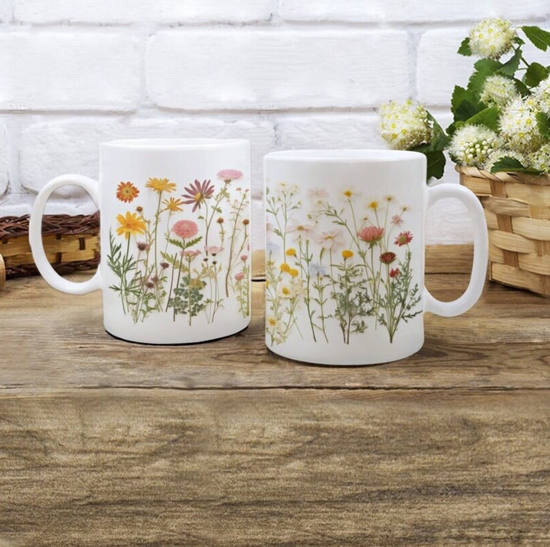 May include: Two white ceramic mugs with pressed flower designs. One mug features a variety of pink, yellow, and orange flowers, while the other mug features white and pink flowers.