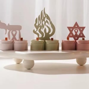May include: A white, wooden decorative candle holder with three sections, each holding two candles. The sections are adorned with cut-out shapes: a pink menorah, a flame, and a Star of David. The candles are in shades of pink and green.