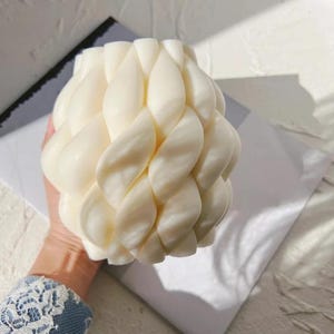 May include: A cream-coloured candle with a woven, braided design. The candle has a rounded shape and is made of a smooth, waxy material. The candle is a decorative item, suitable for home decor or as a gift.