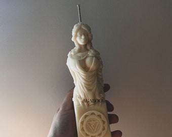 May include: White candle shaped like a woman holding a child. The candle has a symbol of a sun with a triangle inside on the base. The candle is labeled "AGABOO Candles".