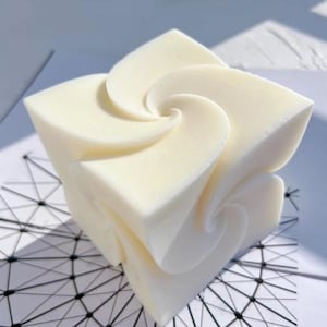 May include: A cream-colored, cube-shaped candle with a unique design. The candle features a series of swirling, petal-like shapes that create a floral appearance. The candle is set against a geometric patterned background.