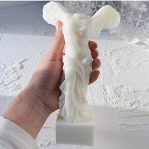 May include: A white, wing-adorned sculpture of a classical figure, standing on a rectangular base. The figure's draped garments and detailed wings are visible. The sculpture is a solid, off-white color.
