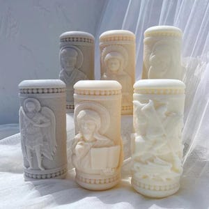 May include: A collection of six off-white, cylindrical religious figurines. Each figurine features detailed relief carvings of religious figures, including angels and saints. The carvings are set against a textured background, and the figurines have a smooth, rounded top.