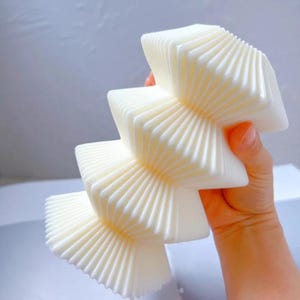 May include: A cream-coloured, accordion-style decorative object with a layered, geometric design. The object is held in a hand and has a textured, pleated surface. The overall shape resembles a stylized star or flower.