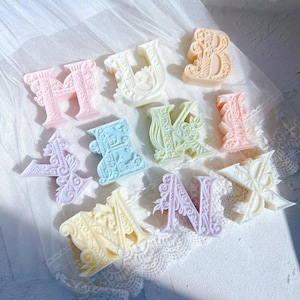 May include: Assortment of pastel-colored decorative letter soaps. The letters include H, U, B, A, E, V, I, M, and K. Each letter has a unique design with floral and ornate details. The soaps are arranged on a white, lace-trimmed surface.