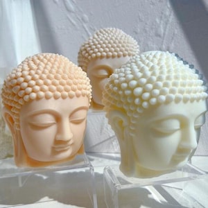 May include: Three Buddha head sculptures in varying shades of cream and off-white. Each head features a serene expression with closed eyes and a textured, bubble-like hairstyle. The sculptures are displayed on clear, rectangular stands.