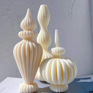 Può includere: Tre candele decorative color avorio con disegni ornati e a coste. Le candele variano nella forma, tra cui un terminale alto e appuntito, una forma sottile e allungata e una forma arrotondata e bulbosa. Sono disposte su una superficie chiara.