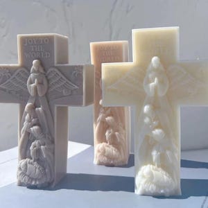 May include: Three cross-shaped candles in varying shades of white and beige. Each candle features a detailed relief of an angel and the text "JOY TO THE WORLD" at the top. The candles are a religious or holiday-themed item.