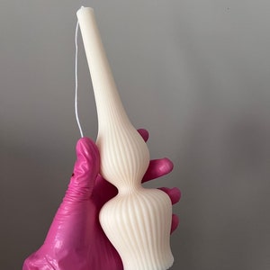 Op de afbeelding: Een crèmekleurige kaars met een unieke, langwerpige vorm. De kaars heeft een geribbelde textuur en een lont die zich van bovenaf uitstrekt. De kaars wordt vastgehouden door een hand met een roze handschoen. De kaars is ongeveer 18 cm hoog.