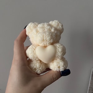 May include: A cream-coloured teddy bear figurine with a heart-shaped accent. The bear has a textured, rose-like pattern and a smooth heart in the centre. The figurine is held in a hand against a neutral background.