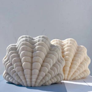 May include: Two seashell-shaped candles. One is a light cream colour, and the other is a soft white. The candles have a textured, layered design, resembling the ridges of a seashell. They are placed on a light blue surface.