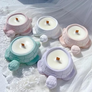 May include: Five turtle-shaped candles in pastel colors: pink, white, blue, green, and lavender. Each candle has a white wax interior and a wooden wick. The turtle shells have a textured, segmented design.