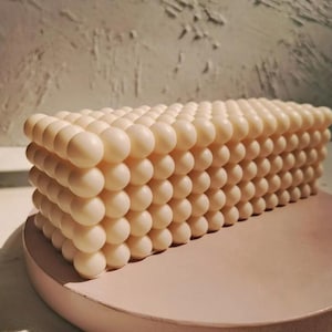 May include: A rectangular, off-white candle with a textured, bubble-like design. The candle is composed of numerous small, spherical shapes arranged in neat rows and columns. It sits on a round, light pink surface, creating a minimalist aesthetic.