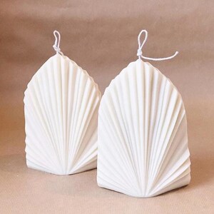 May include: Two white, fan-shaped candles with a textured, ridged design. Each candle has a white wick tied at the top. The candles are set against a neutral background.