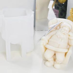 May include: A white silicone mould for a Santa Claus figure and a finished cream-coloured Santa Claus figurine. The Santa Claus figurine has a beard, hat, and a bag of gifts. The mould is a rectangular shape.