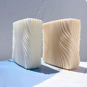 Può includere: Due candele rettangolari con texture a onde. Una è color crema, l'altra beige. Sono posizionate su una superficie azzurra, con sfondo bianco. Le candele misurano circa 10 cm di altezza.