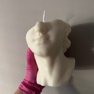 Può includere: Una candela color crema a forma di busto classico. La candela presenta dettagliati tratti del viso, tra cui naso, labbra e mento. Uno stoppino bianco si estende dalla parte superiore della testa. La candela è tenuta da una persona con un guanto rosa.
