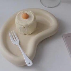 Puede incluir: Una fuente de servir de color crema con forma orgánica con un pequeño pastel blanco cubierto con una guarnición de color melocotón. Un tenedor blanco descansa sobre el plato. El plato está sobre una superficie blanca.