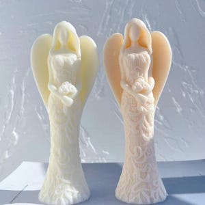 May include: Two white angel figurines with wings, each holding a bouquet of flowers. The angel on the left is wearing a white dress with lace detailing, while the angel on the right is wearing a cream-colored dress with lace detailing.