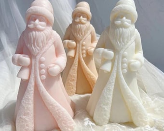 Puede incluir: Tres figuras de Papá Noel en tonos rosa, melocotón y blanco. Cada figura tiene un abrigo largo, un gorro y una barba completa. Las figuras están sobre una superficie de encaje blanco.