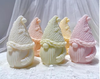 May include: A collection of gnome-shaped soaps in various pastel colors, including white, pink, and yellow. Each soap features a detailed design with a pointed hat, braided beard, and a round body. The soaps are arranged on a white lace surface.