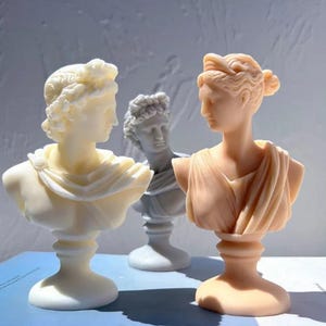 May include: Three classical bust sculptures in varying shades of white and peach. The busts depict figures with detailed hairstyles and draped garments, set against a soft, blurred background. The sculptures are displayed on a light blue surface.
